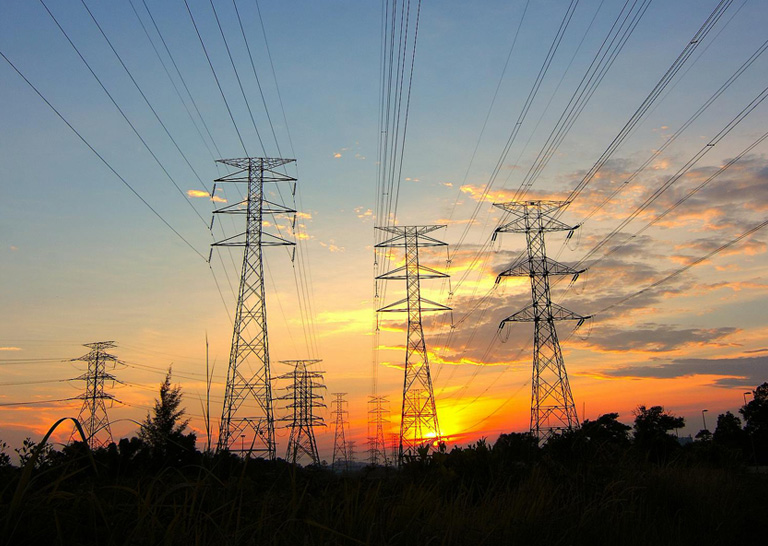 Tall electricity transmission towers stand in silhouette against a colorful sunset sky. Multiple high voltage power lines stretch across the scene, converging toward the horizon. Warm orange and yellow tones contrast with the cool blue clouds, creating a dramatic industrial landscape. Trees and grasses in the foreground add a natural element to the energy infrastructure setting.
