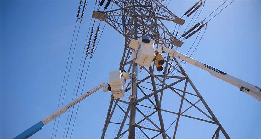 Two bucket cranes elevating toward an electric transmission tower.