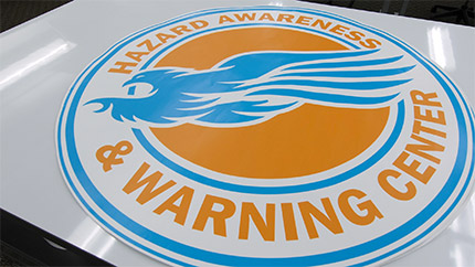 The HAWC logo consisting of an illustration of a blue hawk in the center of an orange disk surrounded by the words Hazard Awareness & Warning Center. A blue outer ring encompasses the entire logo.