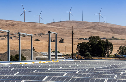 Exterior scene with solar panels in the foreground and windmills on the hills in the distance.