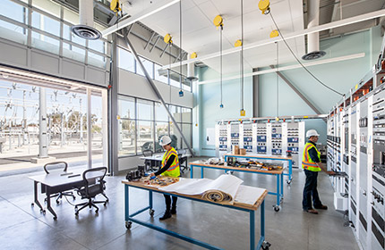 An interior work space of the Electric Safety Academy showing two employees. One is working at a desk with blueprints and hardware components, the other is at a wall of control panels.