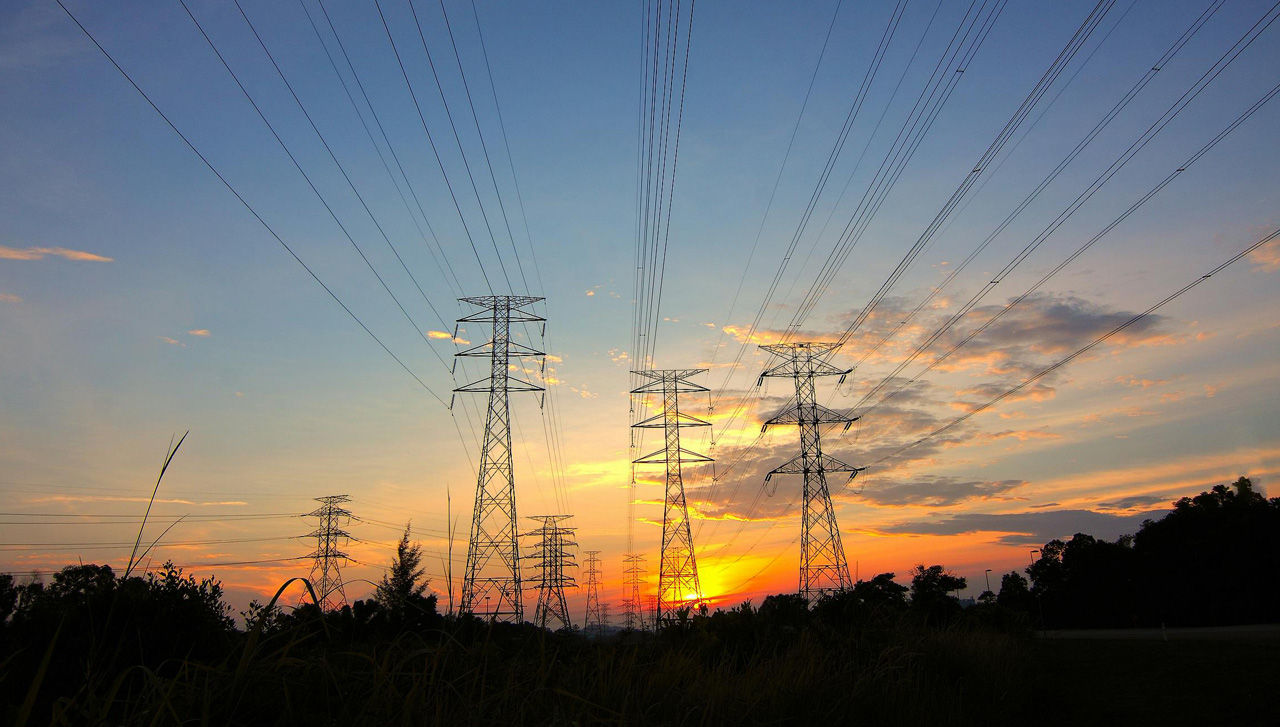 Tall electrical transmission towers stand in a row against a colorful sunset sky. Multiple sets of power lines stretch across the scene, silhouetted by the warm orange and yellow horizon. Soft clouds and fading daylight create a dramatic contrast with the dark structures. Grassy foreground and distant trees frame the industrial landscape.